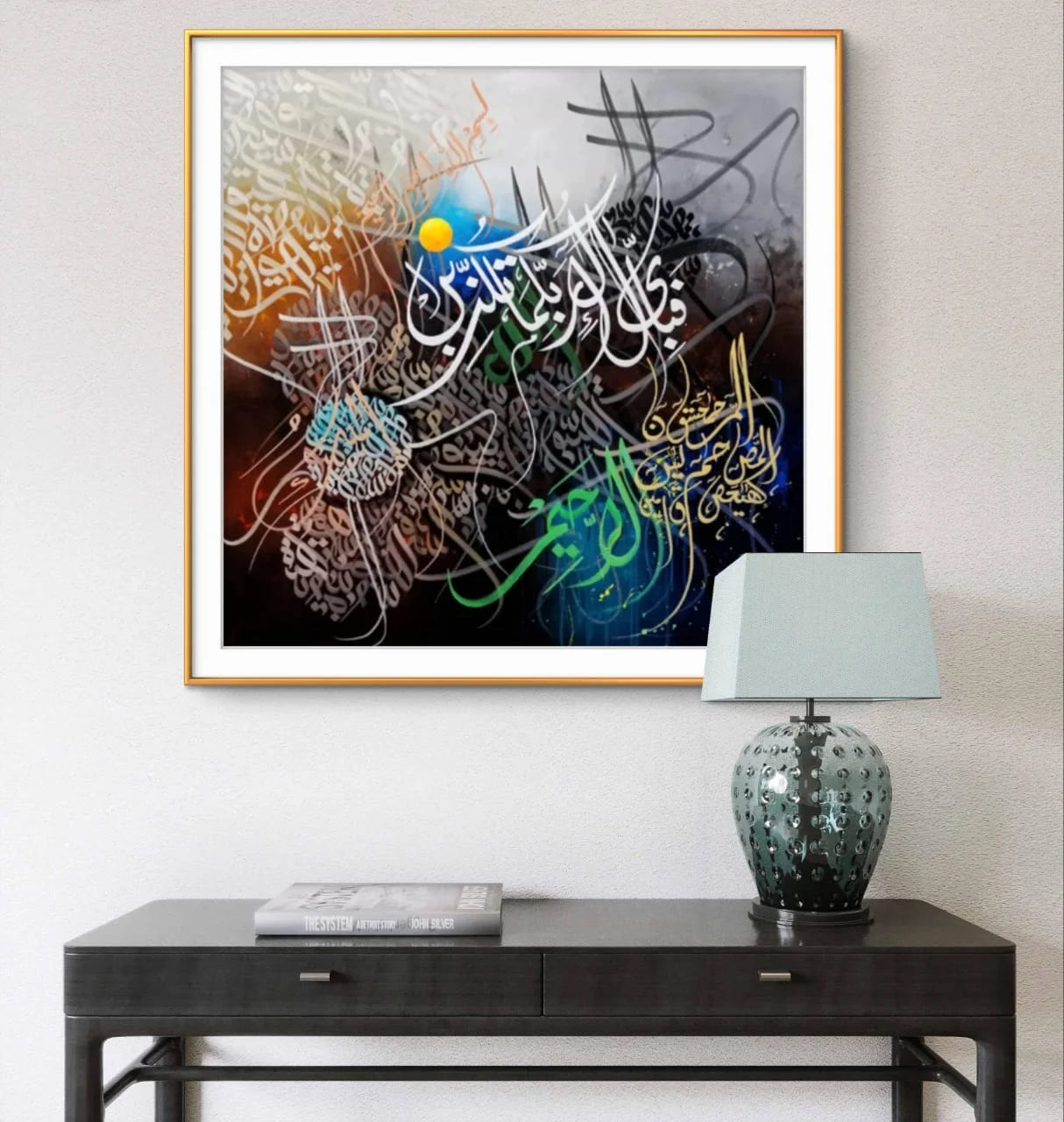 Framed abstract artwork with colorful calligraphy on a wall above a black console table with a lamp.